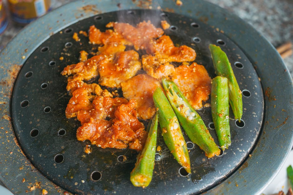 Close-up of marinated meat and okra grilling, emitting steam on an iron skillet.