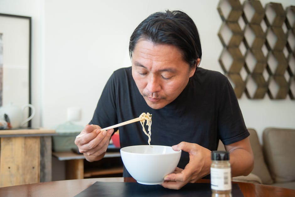 An Asian man eats noodles with chopsticks in a cozy indoor setting, showcasing a homey atmosphere.