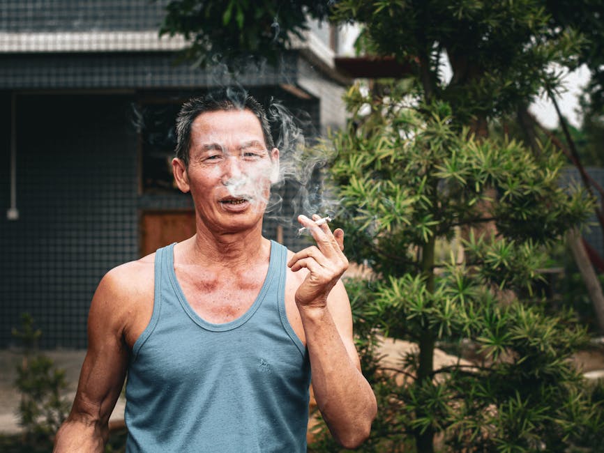 A man in a tank top smokes a cigarette outdoors next to lush greenery.