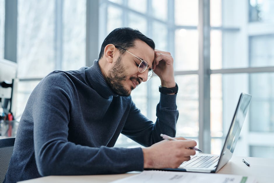 Man looking stressed at laptop in a modern office with large windows.