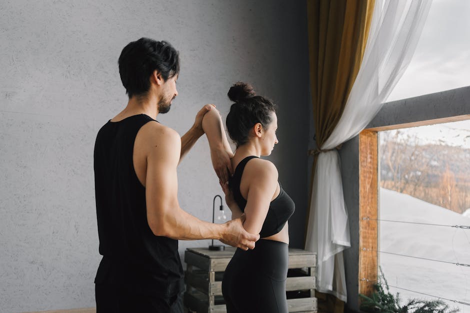 A man and woman practicing partner yoga poses near a large window inside a cozy room.