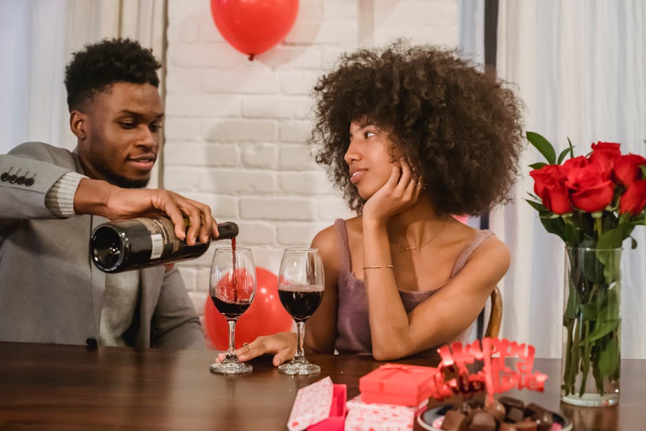 Happy young African American male pouring red wine to girlfriend sitting at table and celebrating Saint Valentine Day