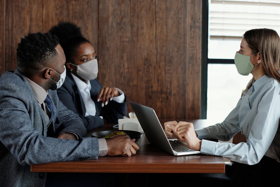 Three coworkers in masks discussing a project indoors with a laptop during the pandemic.