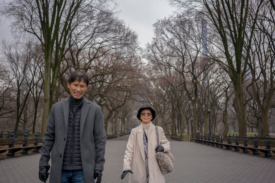 A smiling couple enjoys a leisurely walk in a tree-lined park during a chilly day.