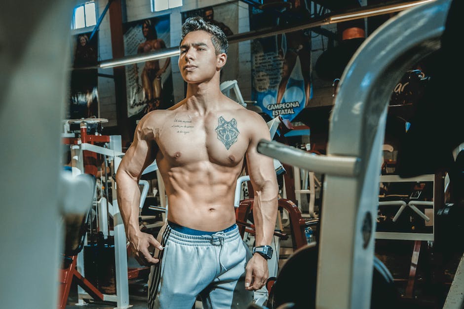 Shirtless man with tattoos posing in a gym environment, showcasing fitness and strength.