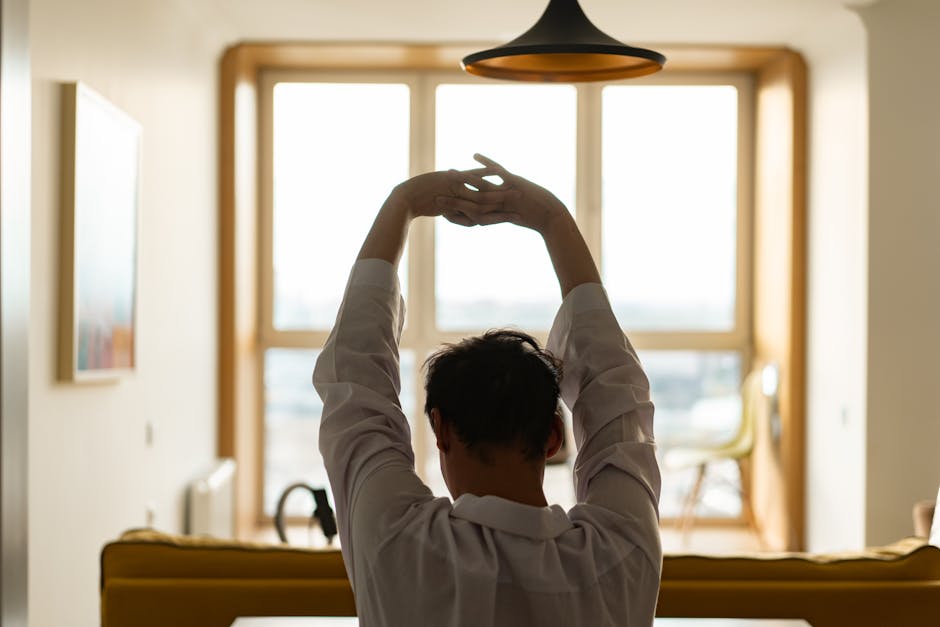 Back view of a man stretching inside a room with a sunny window backdrop.