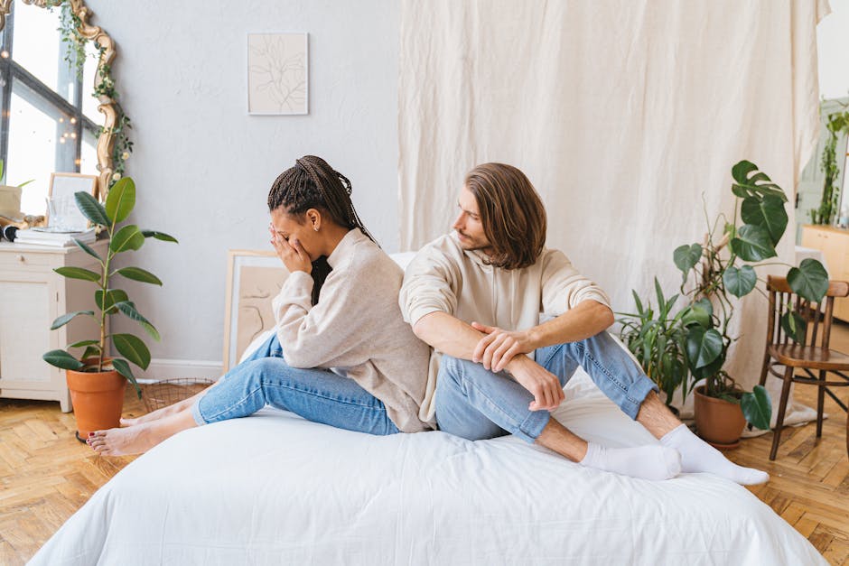 Couple sitting back-to-back on bed, showing relationship conflict in a cozy room.