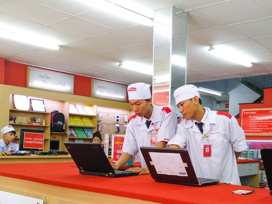 Employees in a tech store assisting at laptops, wearing uniforms, engaging in customer support.
