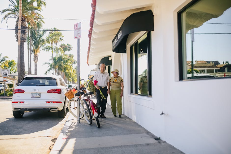 An elderly couple walks hand in hand outside a shop, holding pink flowers, enjoying a sunny day.