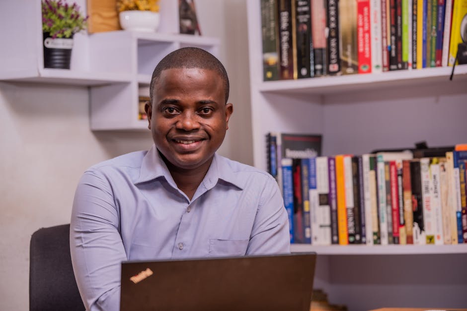 A smiling African American man sitting at a desk in an office with a bookshelf filled with books.