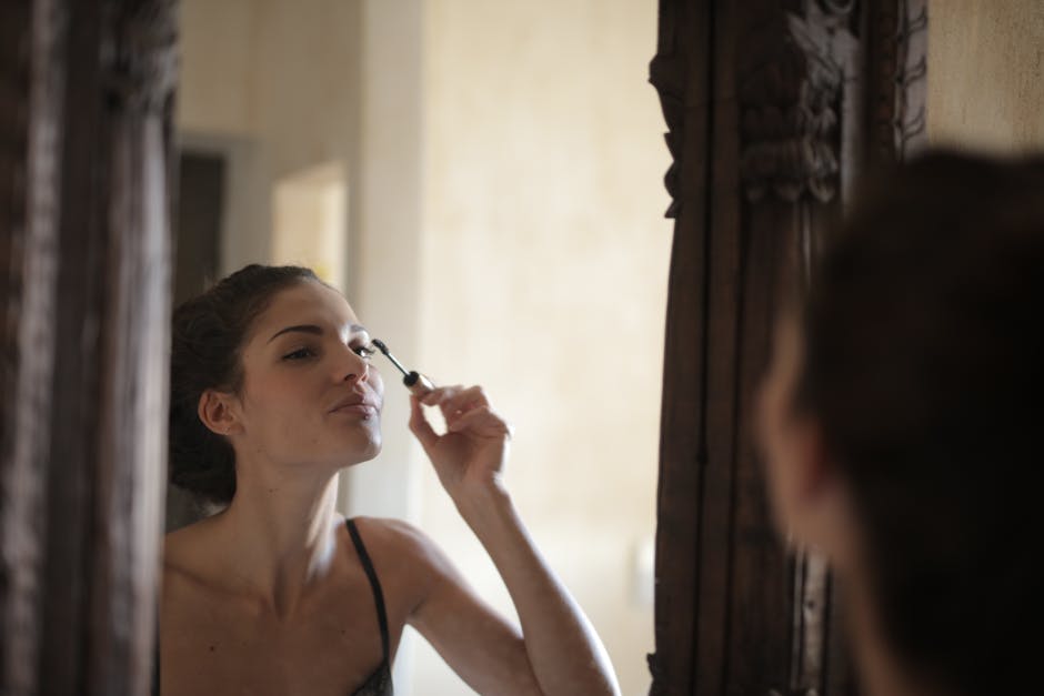 Woman smiling while applying mascara in front of a mirror, reflecting a joyful facial expression.