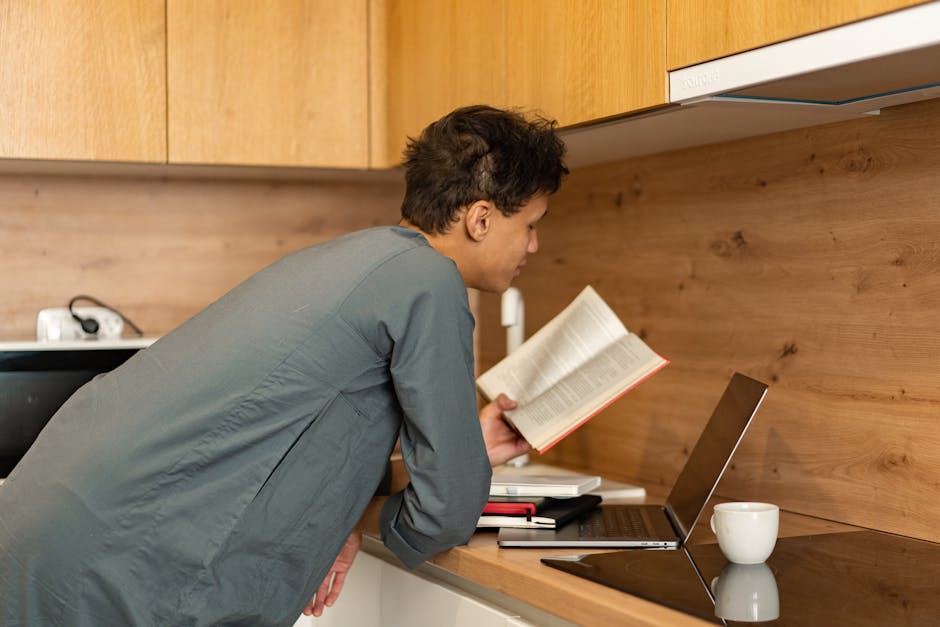 A person reads a book while working on a laptop in a contemporary kitchen setting.