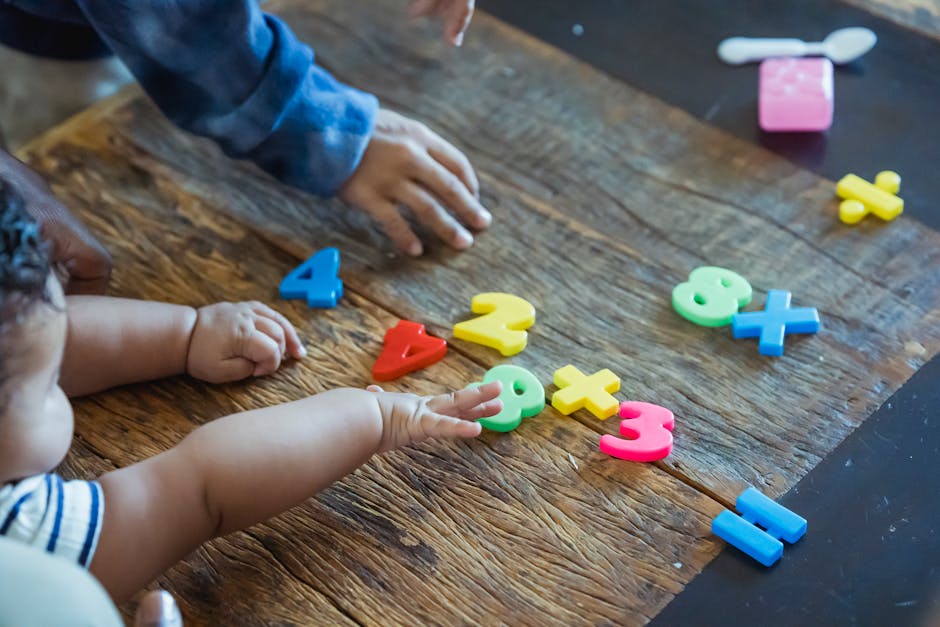 From above of crop anonymous ethnic baby and kid playing with assorted toy numbers and letters at wooden table in house