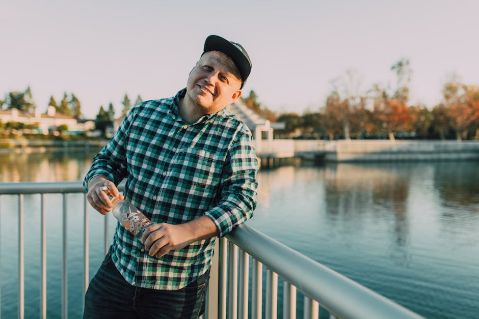 Caucasian man holding bottle casually leaning on metal railing by a river.