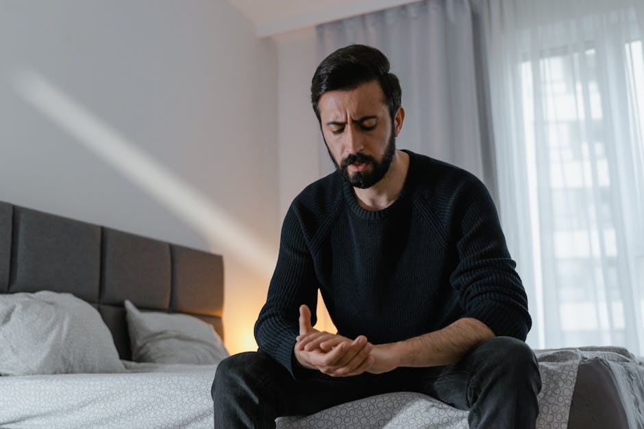 A worried man in a black sweater sits on a bed in a bright room, expressing stress.